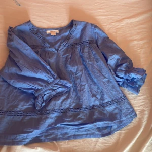 Blå blus med spets från Gina Tricot - Supersöt blå blus från Gina Tricot med v-ringning och fina spetsdetaljer längs halsringningen och ärmarna. Blusen har långa ärmar med resår vid handlederna och en luftig, loose passform. Perfekt för dig som gillar en chill och bohemisk vibe.