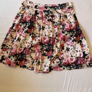 Blommig kortkjol från Dea - Superfin kortkjol från Dea med blommigt mönster i rosa, svart, beige och vitt. Kjolen har veckad design och dragkedja i sidan för enkel påklädning. Perfekt för dig som gillar färg och vill sticka ut lite extra.