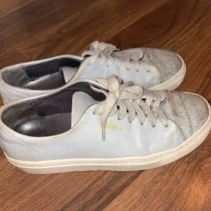 Axel Arigato gråa sneakers med glitter - Snygga gråa sneakers från Axel Arigato med glittriga silverdetaljer på tån. Skorna har klassisk snörning, platt sula och är tillverkade i skinn. Perfekta för dig som vill ha en stilren look med en touch av glitter.