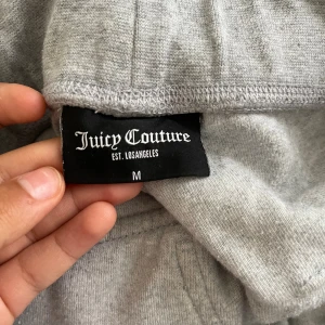 Grå Juicy Couture mjukisbyxor M - Säljer ett par grå mjukisbyxor från Juicy Couture i storlek M. Byxorna har broderad JC-logga på fickan, snörning i midjan och bakfickor med knappar. Perfekta för chill dagar och riktigt sköna tack vare mjukt material.