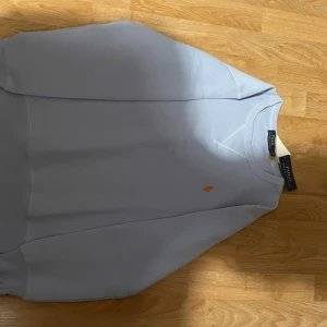 Ljusblå sweatshirt från Polo Ralph Lauren - Ljusblå sweatshirt från Polo Ralph Lauren med klassisk orange broderad logga på bröstet. Tröjan har rund halsringning, långa ärmar och är tillverkad i mjuk bomull. Perfekt för en chill och stilren look.