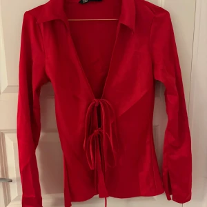 Röd omlottblus med knyt från Zara - Säljer en röd blus från Zara i glansigt material. Blusen har långa ärmar med slits vid ärmslut, djup v-ringning och snygga knytband framtill. Perfekt för dig som vill sticka ut med en färgstark och trendig look.