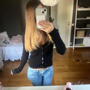 Svart ribbad kofta med knappar - Svart ribbad kofta med v-ringning och långa ärmar. Koftan har en croppad passform och stängs med knappar framtill. Perfekt att slänga över ett linne eller topp för en chill look. Materialet känns mjukt och stretchigt, troligtvis bomull eller en bomullsblandning.