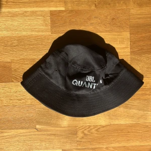 Svart JBL Quantum bucket hat - Bucket hat i svart med JBL Quantum-logga tryckt i vitt framtill. Klassisk form med bred, nedåtvinklad brätte och små ventilationshål på sidorna. Tillverkad i bomullstyg som känns mjukt och slitstarkt. Perfekt för dig som gillar streetwear och vill ha en clean look.