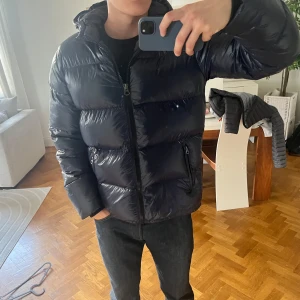 Marine blå pufferjacka med huva från Schott  - Säljer en marinblå pufferjacka från Schott med glansig finish och huva. Jackan har dragkedja framtill, två sidofickor med dragkedja och en clean, modern look. Perfekt för dig som vill ha en varm och stilren jacka till vintern. Ny pris 3000kr