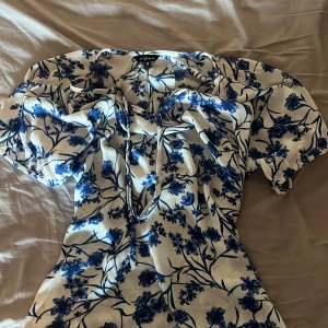 Vit och blå blommig blus amelie & Me - Supersöt blus från amelie & Me i vitt med blått blommigt mönster. Blusen har puffiga korta ärmar, v-ringning med snörning och en lätt figurnära passform. Perfekt för sommarens alla tillfällen och ger en fräsch vibe.