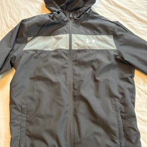 Svart vindjacka från Under Armour - Säljer en svart vindjacka från Under Armour med grått bröstparti och logga. Jackan har huva med snörning, dragkedja framtill och två sidofickor. Perfekt för blåsiga dagar och sportiga outfits.