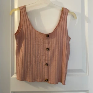 Rosa ribbad linnetopp med knappar - Säljer en söt ljusrosa ribbad linnetopp med knappar framtill. Toppen är ärmlös och har en avslappnad passform, perfekt för varma dagar. Styla med jeans eller kjol för en chill look.