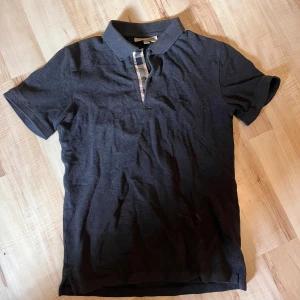 Svart Burberry pikétröja i bomull - Klassisk svart pikétröja från Burberry med korta ärmar och snygg krage. Har den ikoniska rutiga detaljen vid knäppningen och är gjord i mjuk bomull. Perfekt för en clean och stilren look.