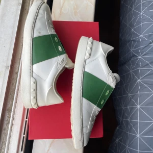 Valentino Garavani Open sneakers vit/grön - Snygga Valentino Garavani Open sneakers i vitt skinn med bred grön rem över mitten. Klassisk rund tå, snörning och ikoniska nitar på sulan. Märkeslogga på plösen och yttersulan. Perfekta för dig som vill ha en clean men ändå unik look. Om du funderar över något skriv så svarar jag, priset är inte hugget i sten vid snabb affär 😊