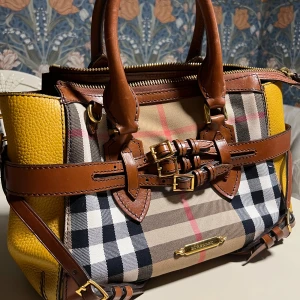 Burberry rutig handväska med läderdetaljer - Snygg handväska från Burberry med klassiskt rutigt mönster, bruna läderdetaljer och spännen i guld. Sidorna är i gul skinn och väskan har både handtag och axelrem. Invändigt finns svart foder och innerficka med dragkedja. Perfekt för dig som vill sticka ut med en ikonisk design.
