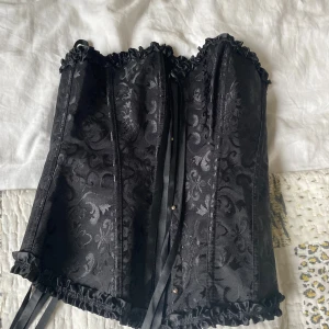 Svart korsett med snörning och mönster - Säljer en svart korsett i storlek M med snyggt blommigt mönster och dekorativa volanger längs kanterna. Korsetten har snörning både fram och bak samt är gjord i polyester med lite stretch. Perfekt för dig som vill ha en edgy och cool look.