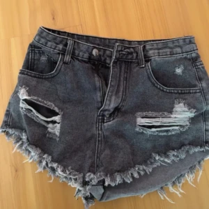 Mörkgrå jeansshorts med slitningar - Snygga mörkgrå jeansshorts med fransig kant och slitna detaljer både fram och bak. Modellen är högmidjad och har klassiska fickor samt dragkedja och knapp. Perfekt för dig som gillar en edgy och avslappnad stil.