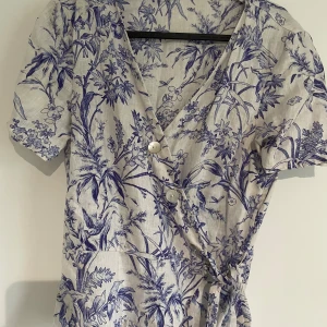 Blommig omlottblus med knyt🤍 - Säljer en kort omlottklänning med vackert blått blommigt mönster på ljus botten. Klänningen har korta ärmar, två pärlemorknappar framtill och ett knyt i midjan. Materialet är linne, vilket gör den superfräsch och luftig. Perfekt för sommarens alla dagar! Köpte den på en second hand butik, vet inte vilken storlek men gissar på S/36🫶🏻