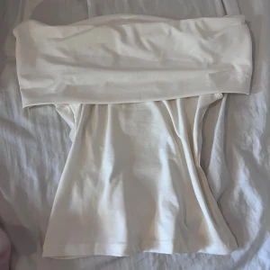 Vit offshoulder topp i stretchigt tyg - Säljer en vit offshoulder topp i mjukt och stretchigt material. Toppen är tight och axelbandslös med en bred kant som vilar över axlarna. Perfekt för dig som gillar en clean och stilren look. Passar snyggt till både jeans och kjol.