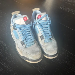 Nike Air Jordan 4 University Blue - Nike Air Jordan 4 University Blue sneakers med ljusblå mocka, meshpaneler och vita samt svarta detaljer. Snygga speckle-detaljer på häl och mellansula, klassisk Jumpman-logga bak och nummer 23 på plösen. Perfekta för dig som vill sticka ut med en ikonisk siluett.