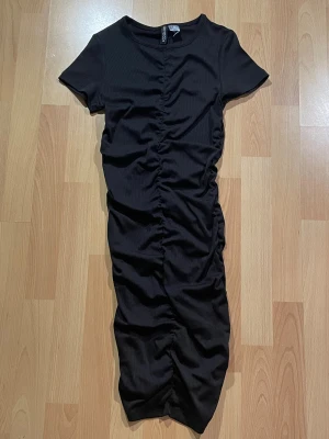 Svart ribbad kortklänning från H&M  - Säljer en svart ribbad kortklänning från H&M i storlek XS. Klänningen har tight passform, korta ärmar och rynkning längs hela fram- och baksidan.