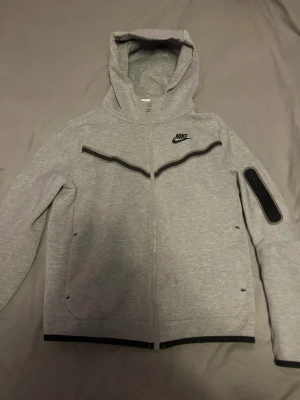 Grå zip-hoodie från Nike - Grå hoodie från Nike med hel dragkedja, svart Nike-logga på bröstet och svarta detaljer längs bröstet och ärmslut. 