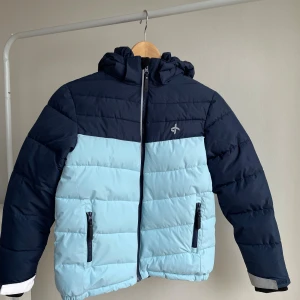 Blå pufferjacka från Cross Sportswear - Snygg pufferjacka från Cross Sportswear i två nyanser av blått. Jackan har avtagbar huva, dragkedja framtill och två fickor med dragkedja. Mjuka muddar vid ärmslut och justerbara kardborreband. Perfekt för kalla dagar och har en sportig vibe. Funkar fint som skidjacka.