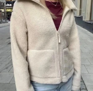 Beige teddyjacka med dragkedja - Mysig beige teddyjacka med hög krage och två stora fickor framtill. Jackan har en rak passform och stängs med dragkedja. Perfekt för att hålla värmen under kyliga dagar och ger en chill vibe till din outfit.