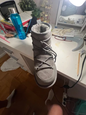 Grå moon boots från Pace - Grå moon boots från Pace med mjuk och fluffig insida i syntetpäls. Skorna har snörning framtill och en rejäl, mönstrad sula för bra grepp. Utsidan är i mockaliknande material och de är perfekta för kalla vinterdagar.