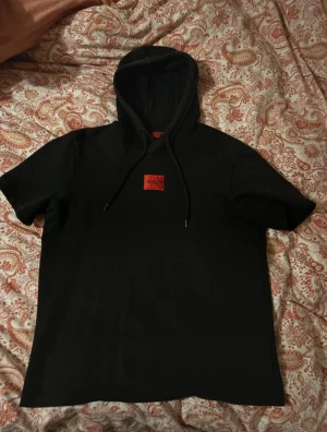 Hugo boss svart hoodie och svart shorts  - Svart hoodie från Hugo Boss med korta ärmar och huva med snörning. Tröjan har en röd Hugo-logga på bröstet och är tillverkad i mjuk bomull. Perfekt för dig som gillar streetwear och vill ha en clean look med en touch av färg. Vill man ha bara hoodie så är det 250kr, vill man ha bara shortsen så är det 200kr o h båda 400kr tillsammans. 