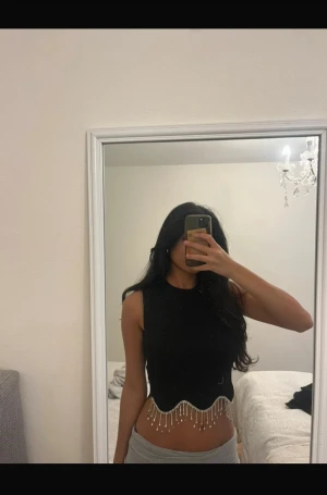 Svart croppad linnetopp med pärlor - Säljer en svart croppad linnetopp med ärmlös design och rund hals. Toppen har vågig nederkant dekorerad med hängande pärlor som ger en extra glittrig vibe. Perfekt för dig som vill sticka ut med coola detaljer och snygg siluett.