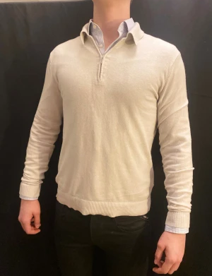 Tiger of sweden tröja  - Snygg beige stickad tröja från Tiger of Sweden i storlek S. Tröjan har krage, half zip och broderad detalj med '1903' vid nederkanten. Ribbad mudd vid ärmslut och nederkant, perfekt för dig som gillar stilrena och klassiska plagg.