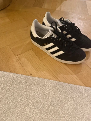 Svarta Adidas Gazelle sneakers - Adidas Gazelle sneakers i svart mocka med vita detaljer och klassiska tre ränder på sidan.  Storlek 38,5. Använda men i bra skick