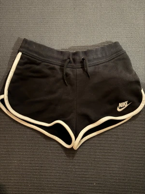 Svarta Nike shorts med vit kant - Nike shorts i svart med vit kant och broderad logga på framsidan. Modellen är loose fit med snörning i midjan och rundade benkanter. Perfekta för träning eller chill, tillverkade i mjuk bomull för skön känsla. Snygg sportig look som funkar året runt.