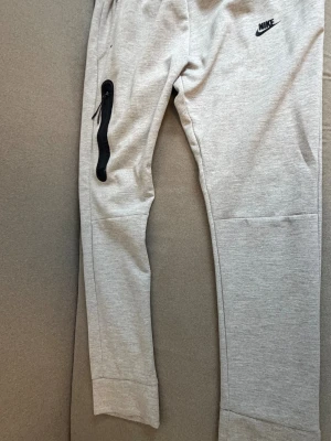 Grå mjukisbyxor från Nike - Snygga grå mjukisbyxor från Nike med svart logga på höften och en cool svart dragkedja längs benet. Byxorna har smal passform och är gjorda i mjukt bomullsmaterial. Perfekta för chill eller träning.