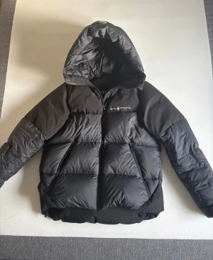 Sail Racing dunjacka  - Säljer en svart dunjacka från Sail Racing med huva och dragkedja. Jackan har en clean look med logga på bröstet, två snedställda fickor (även en innerficka)  med dragkedja och är vadderad med dun från Allied Feather + Down. Perfekt för kalla dagar och riktigt snygg passform. Jackan är i kvinnlig modell men funkar för män också. Köpte jackan för 1 år sen, den är använd under vintern men inga skador och i gott skick. Ursprungligen värd 3500 när jag köpte den!