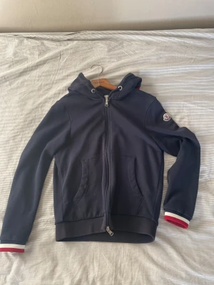 Marinblå Moncler hoodie med dragkedja - Snygg marinblå hoodie från Moncler med huva och hel dragkedja framtill. Tröjan har långa ärmar med ribbade muddar i vitt och rött, samt Moncler-logga på vänster ärm. Praktiska fickor framtill och snörning i huvan. Perfekt för chill dagar.