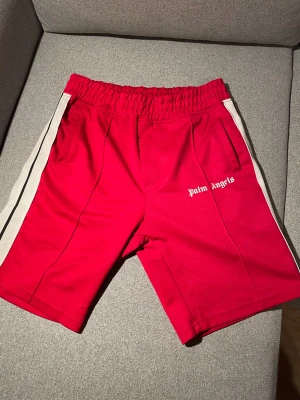Palm Angels röda shorts med vita ränder - Säljer ett par röda Palm Angels shorts med vita och svarta sidoremsor. De har elastisk midja, två fickor fram och Palm Angels-logga tryckt på ena benet. Sportig vibe och perfekt för sommaren. Materialet är mjukt och stretchigt, troligtvis polyester.