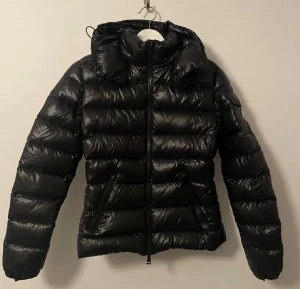Moncler jacka  - Säljer en svart pufferjacka från Moncler med glansig finish och klassisk logga på ärmen. Jackan har huva, dragkedja framtill och två fickor med dragkedja. Dragkedjan framtill och ena dragkedjan till fickan är lite sönder, men de går fortfarande att dra upp utan problem såklart! Perfekt för kalla dagar och riktigt snygg. Köpt på plick så har tyvärr inte något kvitto osv. Size 1 (XS/S)