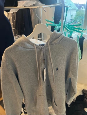 Grå hoodie från Polo Ralph Lauren - Grå hoodie från Polo Ralph Lauren med dragkedja framtill, snörning i huvan och klassisk blå broderad logga på bröstet. Tröjan har långa ärmar, känguruficka och är gjord i mjuk bomull. Perfekt för chill dagar och enkel att matcha med jeans eller joggers.