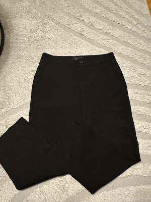 Svarta kostymbyxor - Snygga svarta kostymbyxor med klassisk rak passform och hög midja. Byxorna har knapp och dragkedja framtill samt diskreta fickor bak. Perfekta för en clean och stilren look. Materialet är mjukt och har en lätt stretch.