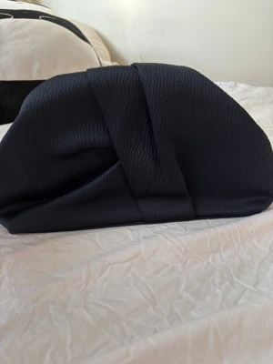 Marinblå clutch från Next - Stilren marinblå kuvertväska från Next med unik omlott-design och struktur i tyget. Väskan har metallram och klassiskt knäppe, samt innerfack med dragkedja för småsaker. Perfekt för dig som gillar minimalistisk och modern look.