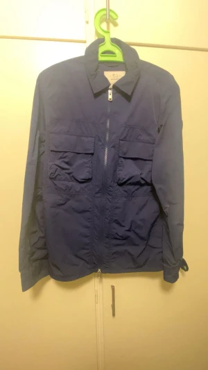 Blå vindjacka från Woolrich, M - Säljer en blå vindjacka från Woolrich i storlek M. Jackan har två stora bröstfickor med lock, dragkedja framtill och klassisk krage. Materialet är lätt och passar perfekt för blåsiga dagar. Diskret Woolrich-logga på ärmen för en clean look.