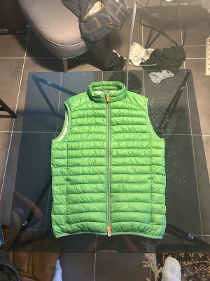 Grön dunväst från Save The Duck - Snygg grön dunväst från Save The Duck med horisontella quiltade paneler och hög krage. Västen har dragkedja med orange detalj och är tillverkad i syntetmaterial med Plumtech-teknologi som är både andningsbar och vattenavvisande. Perfekt för lager-på-lager under kyliga dagar.