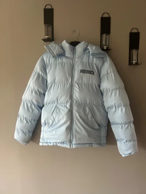 Ljusblå pufferjacka från Hoodrich - Säljer en ljusblå pufferjacka från Hoodrich med stor logga på bröstet och ryggen. Jackan har huva, dragkedja framtill och två snedställda fickor. Insidan har svarta band med H-loggor. Perfekt för kalla dagar när du vill sticka ut.