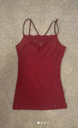Vinröd ribbad linnetopp från Hollister - Supersnygg vinröd linnetopp från Hollister med tunna axelband och spetsdetalj vid halsen. Toppen är ribbad och har en liten metallbricka med Hollister-logga längst ner. Perfekt för dig som gillar stilrena och feminina plagg.