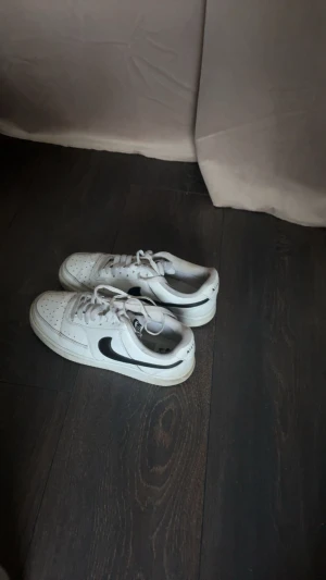 Nike Air Force 1 vita sneakers - Säljer ett par klassiska Nike Air Force 1 sneakers i vitt med svart swoosh. Skorna har snörning, perforerad tå och platt sula. Perfekt för dig som gillar streetstyle och vill ha en tidlös sneaker i garderoben.