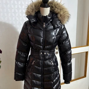 Svart Moncler dunjacka med päls - Snygg svart glansig dunjacka från Moncler med stor huva och lyxig pälsdetalj. Jackan har markerad midja med bälte, dragkedja framtill och två fickor med dragkedja. 