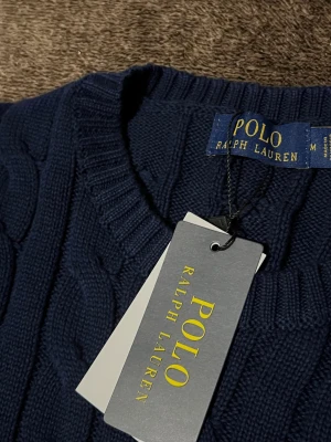 Marinblå stickad tröja Polo Ralph Lauren - Snygg navyblå stickad tröja från Polo Ralph Lauren med klassiskt kabelstickat mönster och röd broderad logga på bröstet. Tröjan har rund hals, ribbade muddar och långa ärmar. Perfekt för dig som gillar stilren och tidlös design.