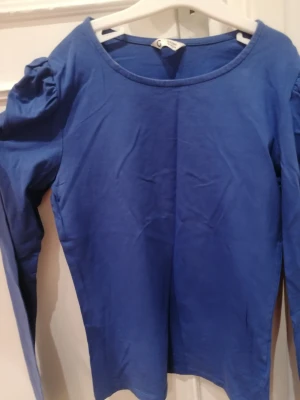 Blå långärmad topp med puffärm - Snygg blå långärmad topp från C i storlek 158/164. Modellen har puffade axlar med rynkade detaljer som ger en cool och trendig look. Tillverkad i mjuk bomull som är skön mot huden. Perfekt för dig som gillar att sticka ut med stilrena detaljer. Använd få gånger. Passar även mig som bär xs. 