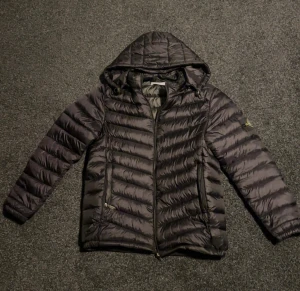 Svart dunjacka från Stone Island - Svart dunjacka från Stone Island med huva och klassisk patch på ärmen. Jackan har quiltad design, dragkedja framtill och två fickor med dragkedja. Perfekt för kalla dagar och riktigt snygg streetstil.