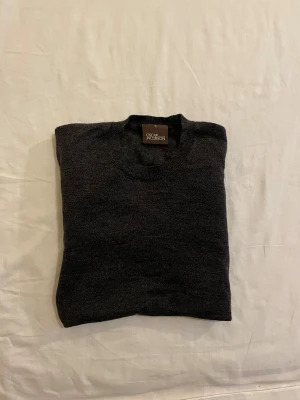 Mörkgrå rundhalsad tröja Oscar Jacobson - Snygg mörkgrå rundhalsad tröja från Oscar Jacobson, modell Custer Roundneck. Tröjan är tillverkad i Italien och har en klassisk, stilren design med ribbade muddar vid ärmslut och nederkant. Perfekt för dig som gillar enkel och tidlös stil.