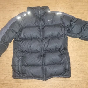 Svart pufferjacka från Nike - Säljer en svart pufferjacka från Nike med reflekterande detaljer på axlarna och dragkedja framtill. Jackan har hög krage och två sidofickor, perfekt för kalla dagar. Klassisk Nike-logga på bröstet och quiltad design för extra värme.