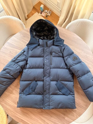 Mörkblå dunjacka Moose Knuckles - Snygg mörkblå dunjacka från Moose Knuckles med avtagbar huva, dragkedja och tryckknappar. Jackan har flera fickor, logga på ärmen och quiltad design. Perfekt för kalla dagar och riktigt street i stilen. Jackan är väldigt varm så du kommer aldrig att frysa. Jackan är köpt på Johnells förra året, har även kvar kvittot. Använd fåtal gånger och ser så gott som ny ut! Pris kan diskuteras!